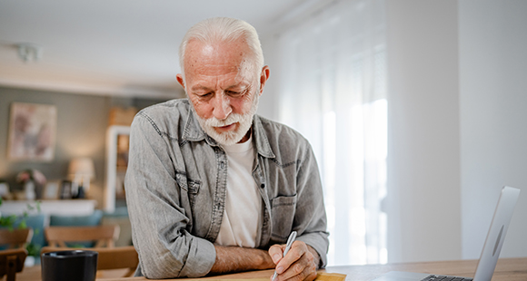 an elderly man writing on a sheet of paper in front of a laptop 